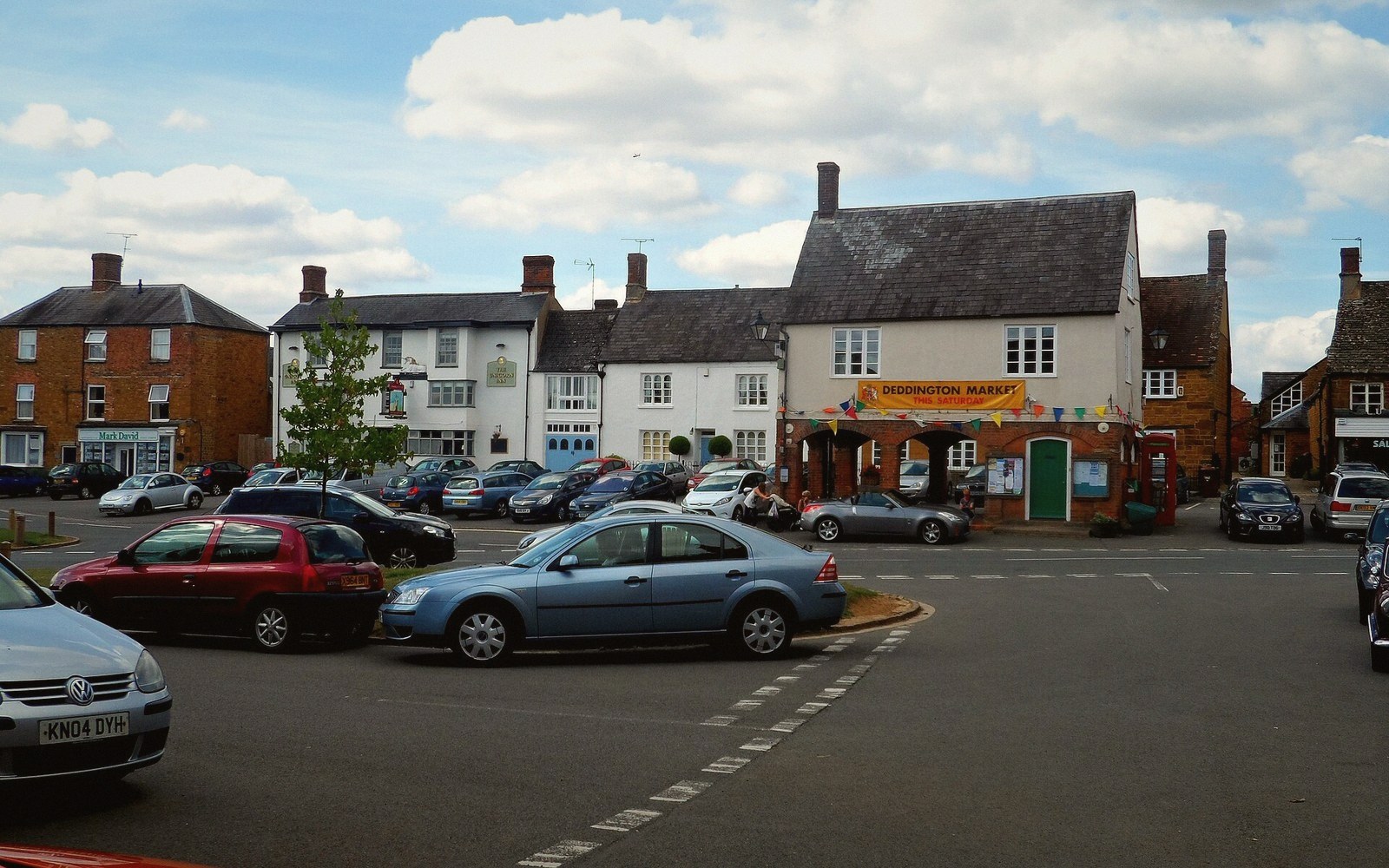 Cars parked in Deddington's market square lined with brick and stone buildings