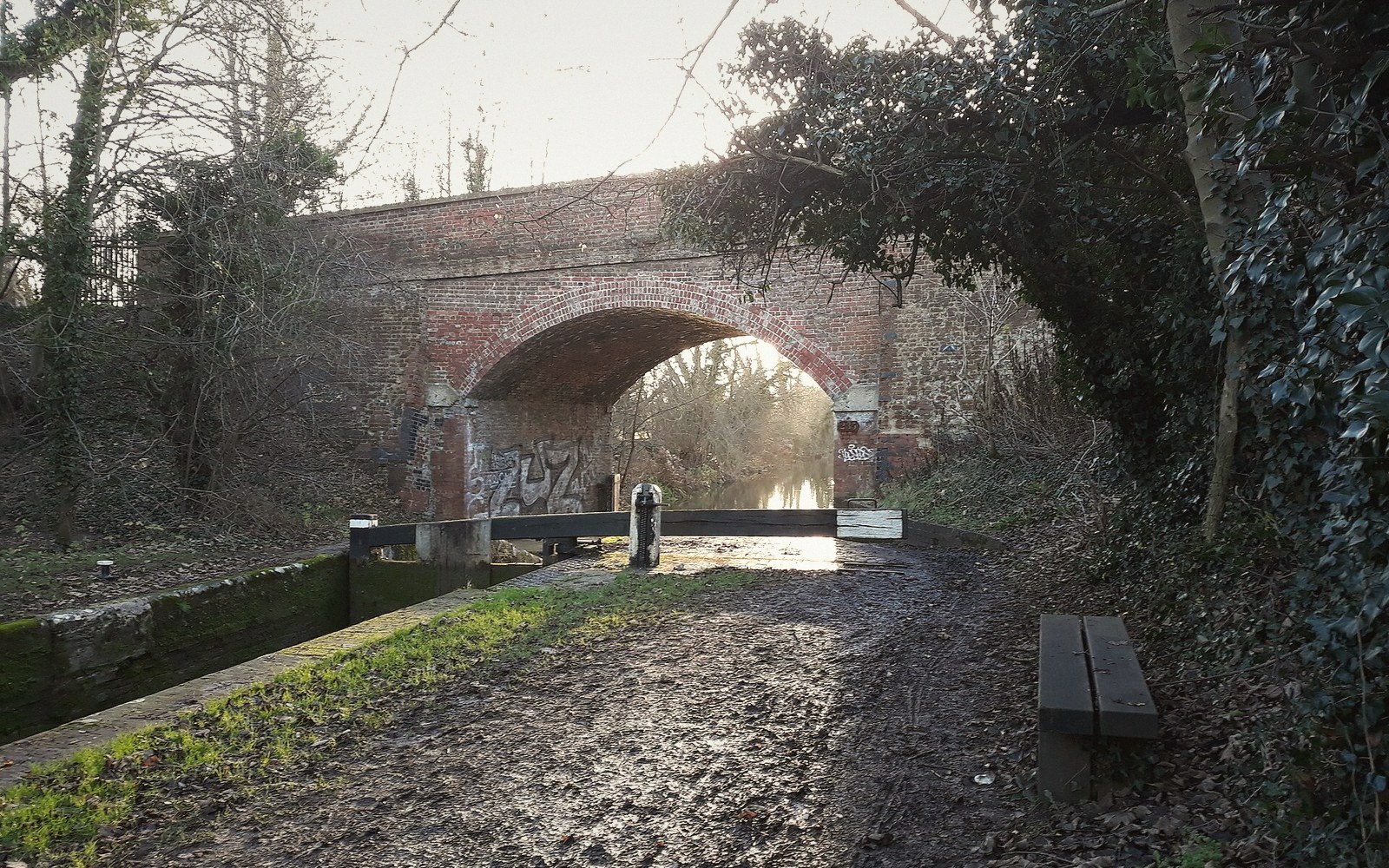 Brick arch bridge over a canal lock with a muddy towpath and bench