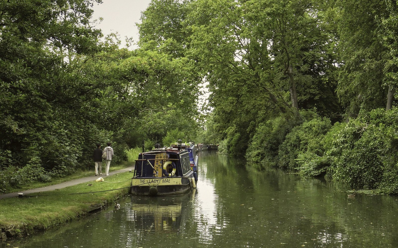 Narrowboat moored along a tree-lined canal towpath with two people walking nearby